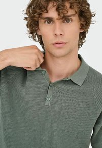Grøn textured polo skjorte med knap-krave, der har et lille mønster og raglan ærmer, båret af en person med krøllet hår.