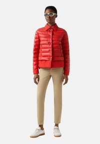 Rote Steppjacke mit Knopfverschluss und Kragen, kombiniert mit beigen Hosen und weißen Turnschuhen. Modell trägt runde Sonnenbrillen.