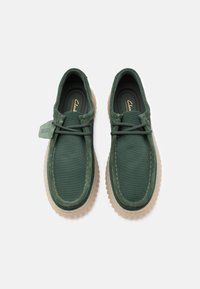 Ζευγάρι από σκούρα πράσινα casual παπούτσια από σούεντ και ύφασμα με μπεζ λαστιχένιες σόλες και κορδόνια, εμφανιζόμενα από πάνω σε λευκό φόντο.