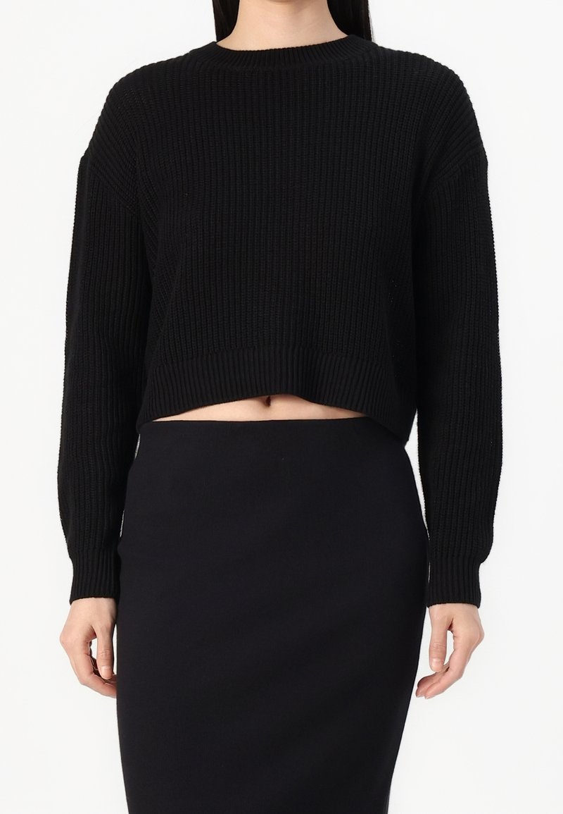 Femme portant un pull court en maille côtelée noire à manches longues et une jupe noire taille haute, debout sur un fond blanc.