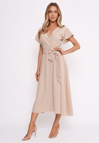 Beiges Wickelkleid mit fließenden Ärmeln, selbstbindendem Taillendetail und Midi-Länge. Glatte Stoffoberfläche und schlichtes Design. Ergänzt durch nudefarbene Absatzschuhe.