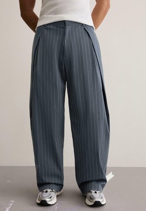 Osoba nosząca luźne, szare spodnie w drobną prążek i białe trampki, stojąca z rękami za plecami na jednolitym tle.