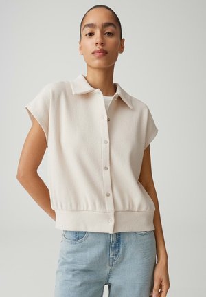 Donna con capelli raccolti, che indossa un top crema senza maniche con bottoni e jeans azzurri, in piedi davanti a uno sfondo chiaro e uniforme.