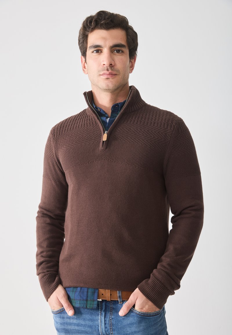 Uomo che indossa un maglione a collo zip marrone sopra una camicia a quadri blu, con le mani nelle tasche dei jeans, in piedi contro uno sfondo bianco uniforme.