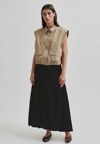 Beige, mouwloze, knoopsluiting vest met voorkruiszakken, gecombineerd met een lange, zwarte, vloeiende rok. De outfit heeft een minimalistisch ontwerp en soepele texturen.