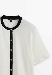 Chemise blanche à manches courtes avec col rond noir et bordure noire verticale ornée de six boutons décoratifs dorés sur le devant.