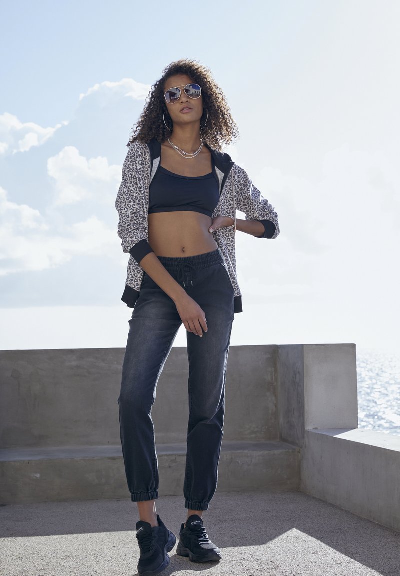 Schwarzes Crop-Top, gemusterter grauer Reißverschluss-Hoodie, dunkle Jogginghose, schwarze Sneakers, große Sonnenbrille, stehend vor einem hellen Himmel.