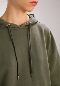 Felpa verde oliva con una grande tasca frontale, cappuccio regolabile con cordini e asole metalliche, realizzata in morbido tessuto di cotone testurizzato.