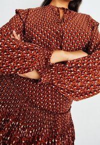 Rotes gemustertes Kleid mit langen, gerafften Ärmeln und einem gesmokten Taillenbund. Verfügt über geometrische und kreisförmige Designs in Weiß und Orange.