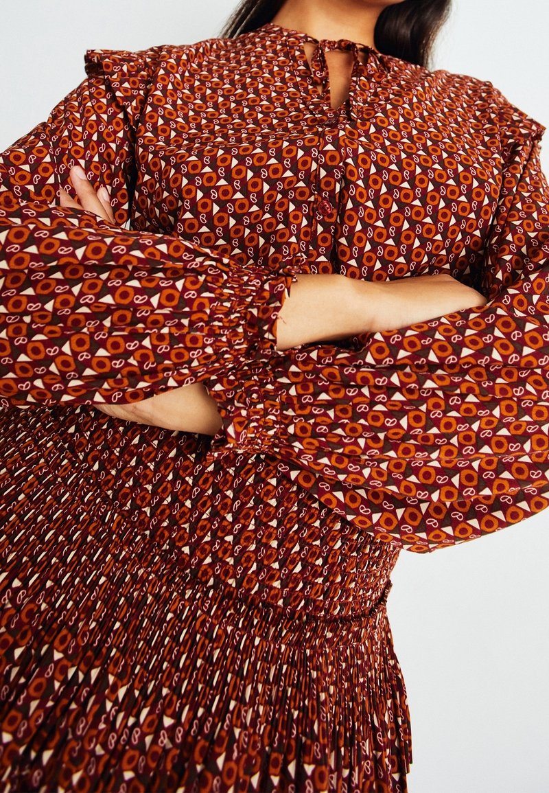 Rotes gemustertes Kleid mit langen, gerafften Ärmeln und einem gesmokten Taillenbund. Verfügt über geometrische und kreisförmige Designs in Weiß und Orange.