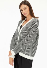 Czarny i biały kardigan w wzór rybiej ości z szerokimi rękawami, głębokim dekoltem w serek oraz kontrastującymi białymi ściągaczami na rękawach i u dołu.