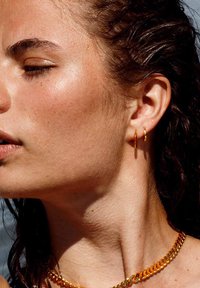 Guld hoop øreringe og en guld kædedyse på en person med vådt, mørkt hår. Huden har et glat, solkysset udseende med skønhedspletter.