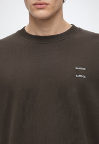 Homme portant un sweat-shirt col rond marron foncé avec du texte blanc "SAMSØE" imprimé deux fois sur la poitrine gauche.
