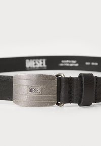 Ceinture en cuir noir avec une boucle en métal argenté texturée, portant le nom de la marque "DIESEL" gravé et une finition lisse dans l'ensemble.