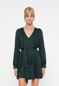 Dark green dress with a V-neckline, long sleeves, and a cinched waist tie. Features a tiered skirt and soft, textured fabric.