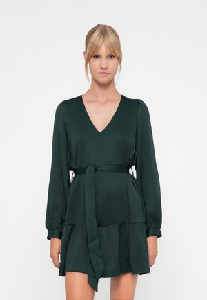 Robe vert foncé avec un décolleté en V, des manches longues et une ceinture cintrée à la taille. Dotée d'une jupe à volants et d'un tissu doux et texturé.