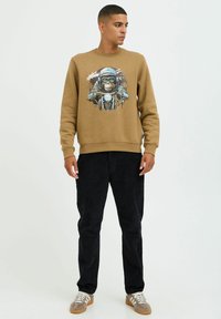 Brun sweatshirt med grafik af en abe iført hjelm, parret med sorte fløjlspants og lysebrune sneakers.