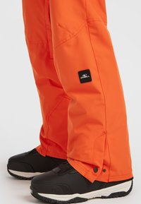 Heldere oranje waterdichte sneeuwbroek met verstelbare drukknopen aan de pijpen, versterkte stiksels en een klein zwart logo-patch op de dij. Zwarte sneeuwlaarzen.