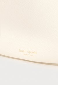 Valge tekstuuriga nahk kuldse kulplommitud "kate spade NEW YORK" logoga. Sile pind õrna mustriga, lisatarvikuid ei ole nähtaval.