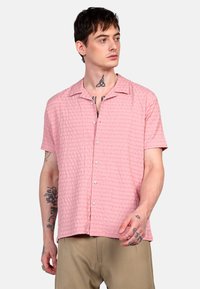 Camisa de botón de manga corta en color rosa con un patrón de cuadrícula texturizado, hecha de tejido ligero. Presenta un cuello con muesca y botones visibles.