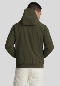 Oliven grønn hettejakke med en tettsittende design, teksturert stoff, ribbede mansjetter og midje, sett fra baksiden.