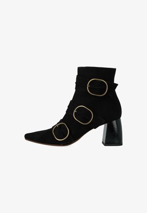Stivale alla caviglia in suede nero con punta affusolata, tre cinturini decorativi con grandi fibbie metallizzate e un tacco spesso e lucido di colore nero.