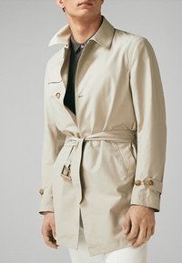 Beżowy trencz wykonany z gładkiego, lekkiego materiału. Posiada paskowany talia, guziki przy mankietach oraz klasyczny kołnierz. Brak wyraźnych wzorów.