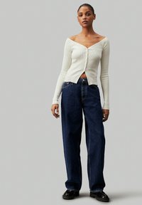 Top blanco de tira acanalada y sin hombros con detalles de botones, combinado con unos vaqueros de denim azul oscuro y suéteres gruesos negros.