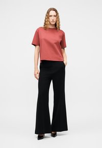 Femme aux cheveux ondulés portant un haut à manches courtes couleur rouille, un pantalon noir à jambes larges et des talons noirs, debout devant un fond uni.