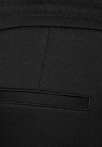 Tela negra con una superficie ligeramente texturizada, que presenta un bolsillo trasero con una solapa plana y rectangular y detalles de costura en la parte superior.