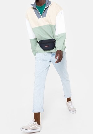 Värilohkoinen svetaritakki beigest, vihreästä ja valkoisesta; kevyt kangas. Päällä vaaleansiniset farkut ja harmaat lenkkarit. Laivastonsininen vyölaukku.