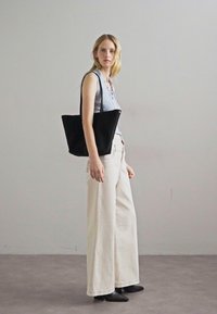 Borsa tote nera realizzata in tessuto morbido, con un design minimalista e due manici. Abbinata a un top senza maniche blu chiaro e pantaloni larghi beige.