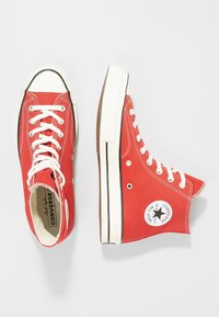 Red canvas high-top sneakers with white laces and rubber soles. Features a black star logo on the side and contrasting stitching details.