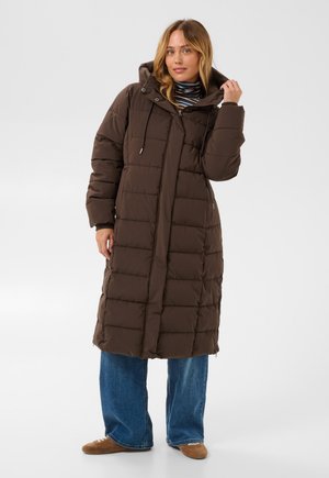 Femme portant un long manteau matelassé marron avec une capuche, un col roulé rayé, un jean bleu et des chaussures marron, debout sur un fond uni.