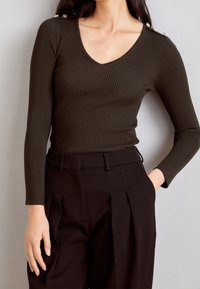Mujer con un top de manga larga de ribete V en marrón oscuro con botones dorados en el hombro y pantalones negros de cintura alta con pliegues.