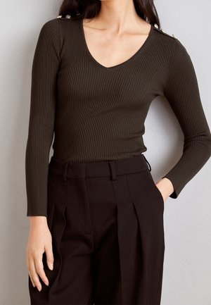 Mujer con un top de manga larga de ribete V en marrón oscuro con botones dorados en el hombro y pantalones negros de cintura alta con pliegues.