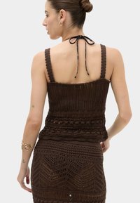 Mujer con el pelo recogido en un moño, vistiendo un top sin mangas de punto texturizado marrón oscuro y falda a juego, con tirantes halter atados en la espalda y pequeños tatuajes en los brazos.