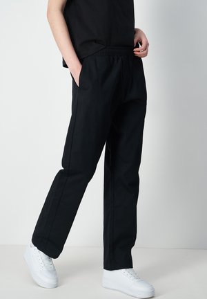 Persona che indossa pantaloni neri con elastico in vita, con le mani nelle tasche e sneaker bianche, in piedi su uno sfondo chiaro e uniforme.