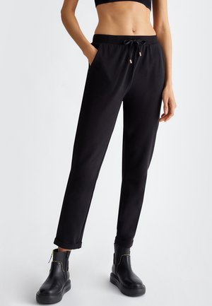 Kobieta ubrana w czarne spodnie ze ściągaczami, podwiniętymi mankietami, czarne botki za kostkę oraz krótki czarny top, stojąca na tle o neutralnym kolorze.