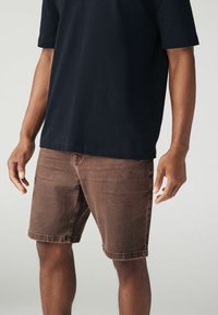 Bărbat îmbrăcat în pantaloni scurți din denim de culoare maro închis și o cămașă neagră cu mânecă scurtă, stând cu brațele relaxate pe lângă corp, pe un fundal simplu.