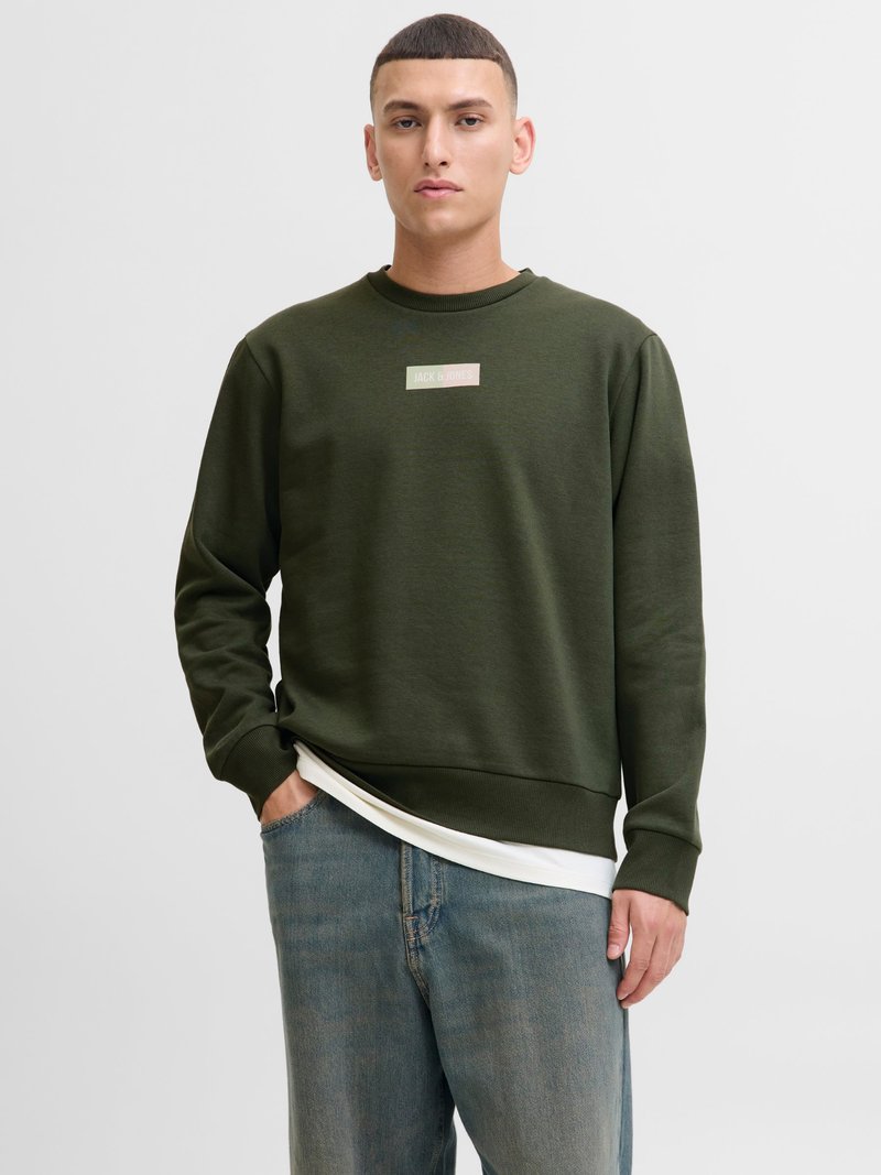 Ung mand iført en olivengrøn sweatshirt med "Jack & Jones"-logo, en hvid skjorte, der stikker frem, og slidte blå jeans, stående foran en ensfarvet baggrund.