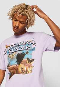 Tricou scurt cu mânecă, de culoare lila, cu un print grafic ce include textul "ZILE ÎNAINTE DE VARĂ" și ilustrații colorate cu fluturi. Textură moale din bumbac.