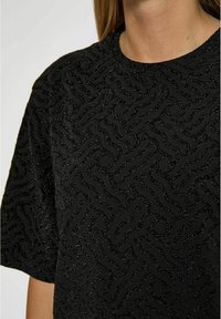 Juodas tekstūruotas viršus su iškilusiu bangų raštu ir metaliniais akcentais, apvaliu iškirpimu ir trumomis rankovėmis. Audinys turi švelnų, audinį primenantį apdailą.