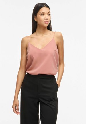 Donna che indossa una blusa rosa polvere senza maniche con spalline sottili e pantaloni neri a vita alta, guarda a sinistra con una mano in tasca.