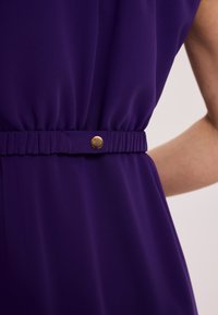 Fioletowa sukienka z marszczonym pasem, akcentowana złotym guzikiem, wykonana z miękkiej, zwiewnej tkaniny o gładkiej fakturze.