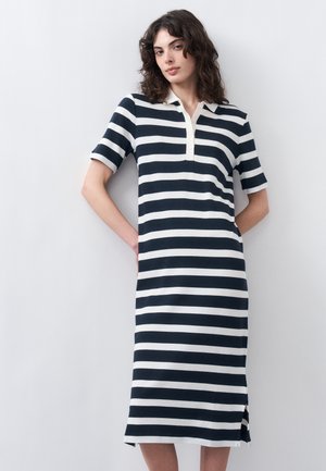 Woman standing against a white wall wearing a knee-length black and white striped polo dress with short sleeves and side slits.