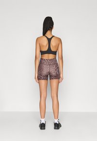 Καφέ αθλητικό σουτιέν και σορτς με λεοπάρ σχέδιο και λεία υφή. Μαύρα αθλητικά παπούτσια, ορατές λευκές κάλτσες μέχρι τον αστράγαλο. Πλάγια όψη από πίσω.