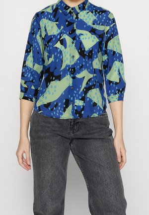 Mujer con camisa de botones de patrón abstracto en azul, negro y verde, con mangas remangadas y vaqueros gris oscuro.