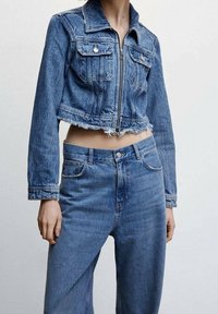 Henkilö, joka on pukeutunut lyhennettyyn siniseen farkkutakkiin, jonka helma on frayed, sekä korkeavyötäröisiin sinisiin farkkuihin, taustan ollessa yksinkertainen.