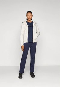 Γυναίκα που στέκει με το ένα χέρι στην τσέπη, φορώντας λευκή φούτερ με φερμουάρ, ναυτικό μπλουζάκι με λογότυπο, ναυτικά παντελόνια και μαύρα παπούτσια σε λευκό φόντο.